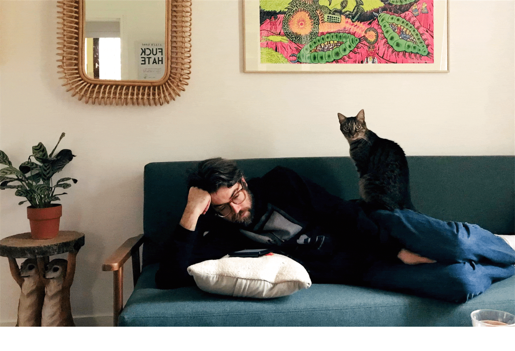 The author Zachary Dillon reclines on a green couch in a living room, reading an e-reader. There is a dark, stripey cat sitting on him and staring at the camera.