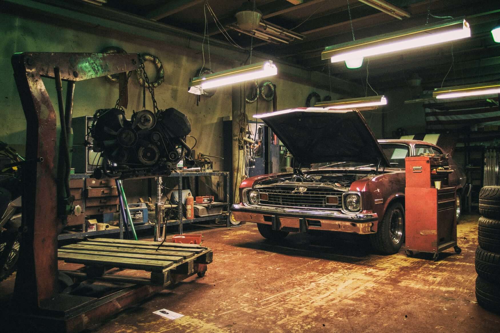 car with hood open and crane holding engine in auto garage at night with fluorescent lights