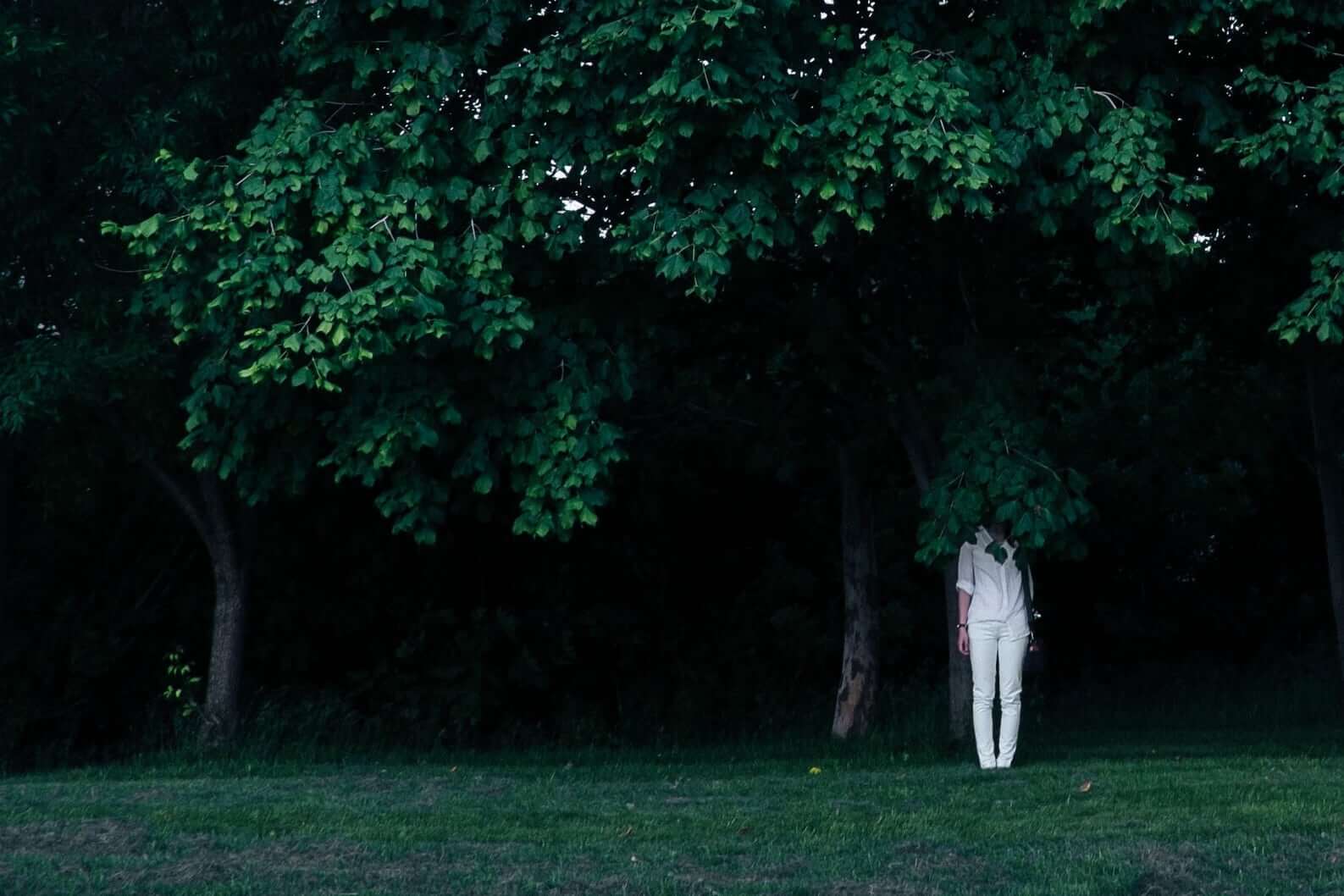 Dark leafy trees, and woman in white shirt and pants standing with face obscured by leaves.