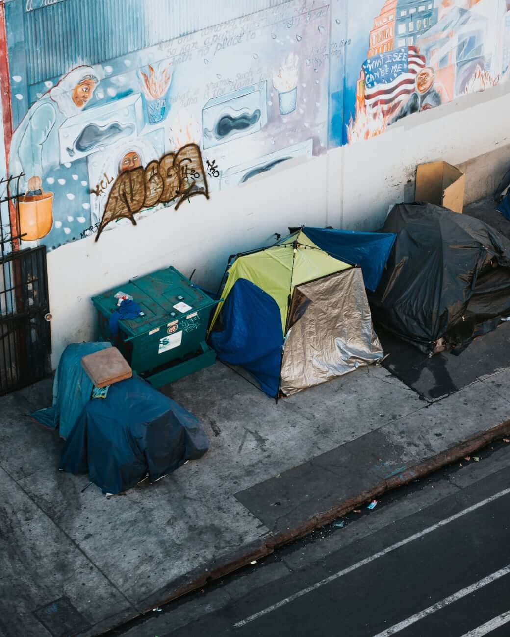 Photo of homeless tent encampment and dumpster on dirty sidewalk in front of a faded mural of an immigrant woman and an American flag