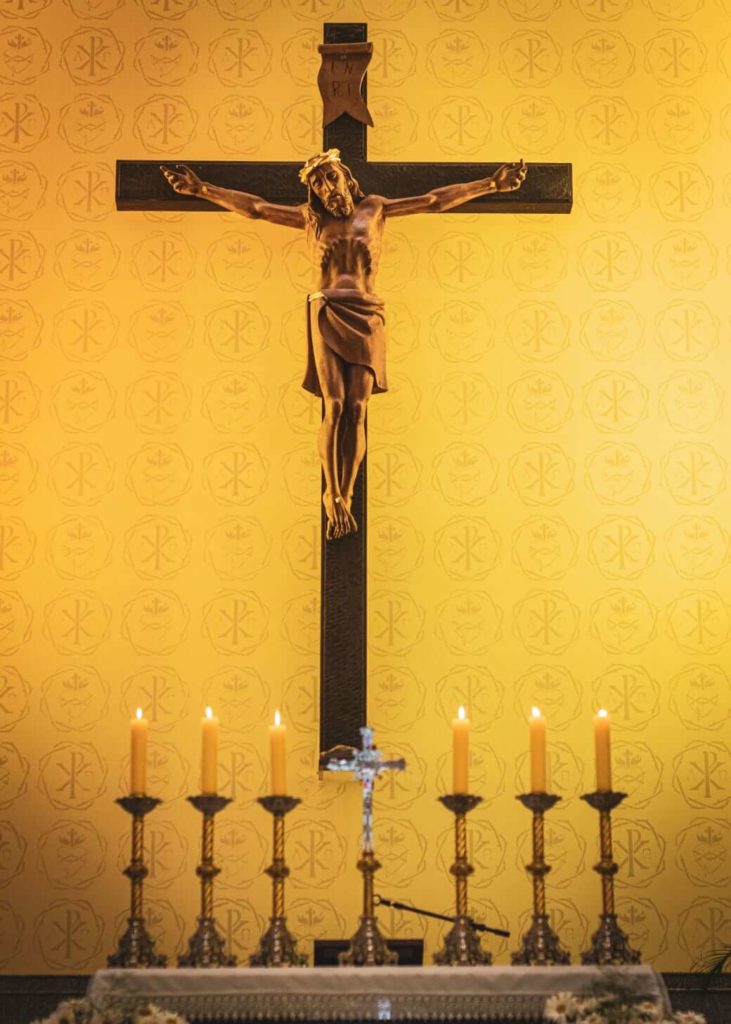 Photo of statuette of Jesus on the cross hanging behind a row of lit candles, with a background of gold wallpaper